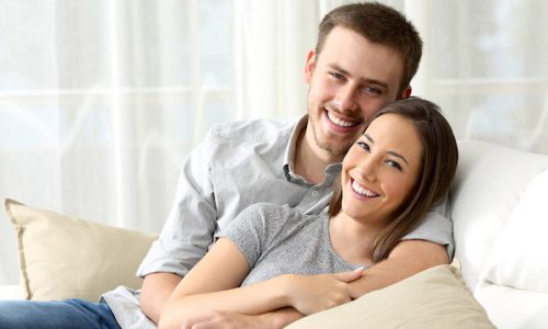 Happy couple sitting in a comfortable couch in the living room and looking at you at home; Shutterstock ID 454334446; PO: Allianz