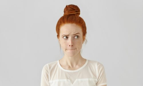 Studio isolated shot of doubting confused cute young redhead freckled female looking sideways, pursuing or pinching her lips, feeling uncertain and indetermined about exposing important information