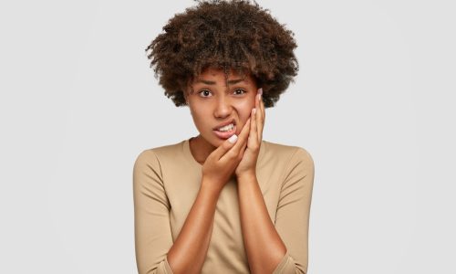 Isolated shot of displeased dark skinned young female has Afro haircut, keeps hand on cheek, frowns face from pain, suffers from toothache, dressed in casual sweater, expresses negative emotions